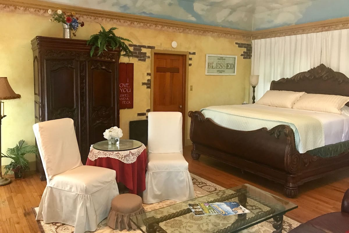 A bedroom with a bed, two chairs, a small table with a vase, a red sign, and a ceiling mural.