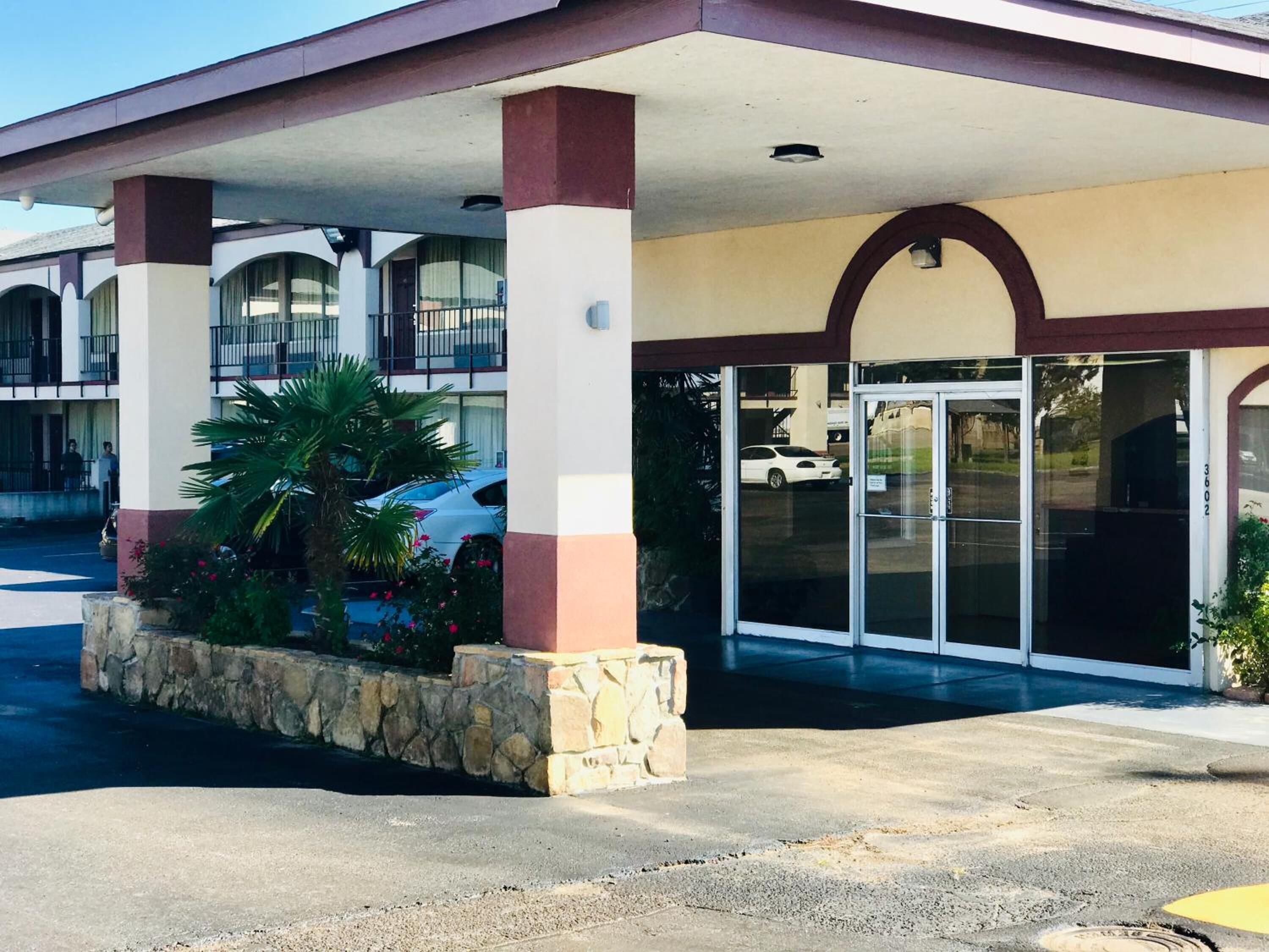 Ein Hotel mit Eingangsbereich und überdachtem Parkplatz, ein Steinpflanzgefäß mit Bepflanzung sowie ein Auto im Hintergrund.