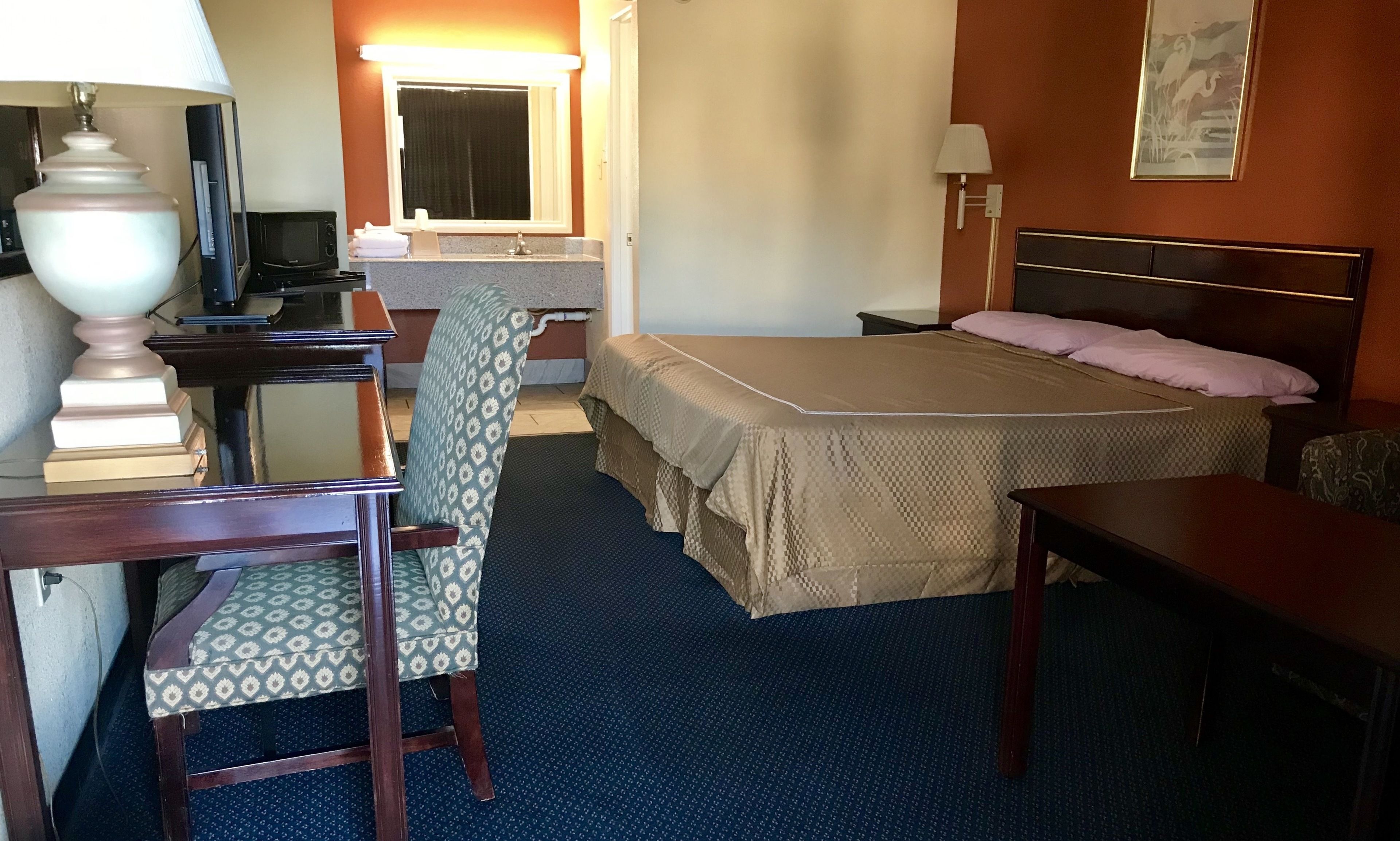 Habitación de hotel con cama, escritorio con lámpara, silla, pequeña mesa, espejo y un cuadro en la pared.