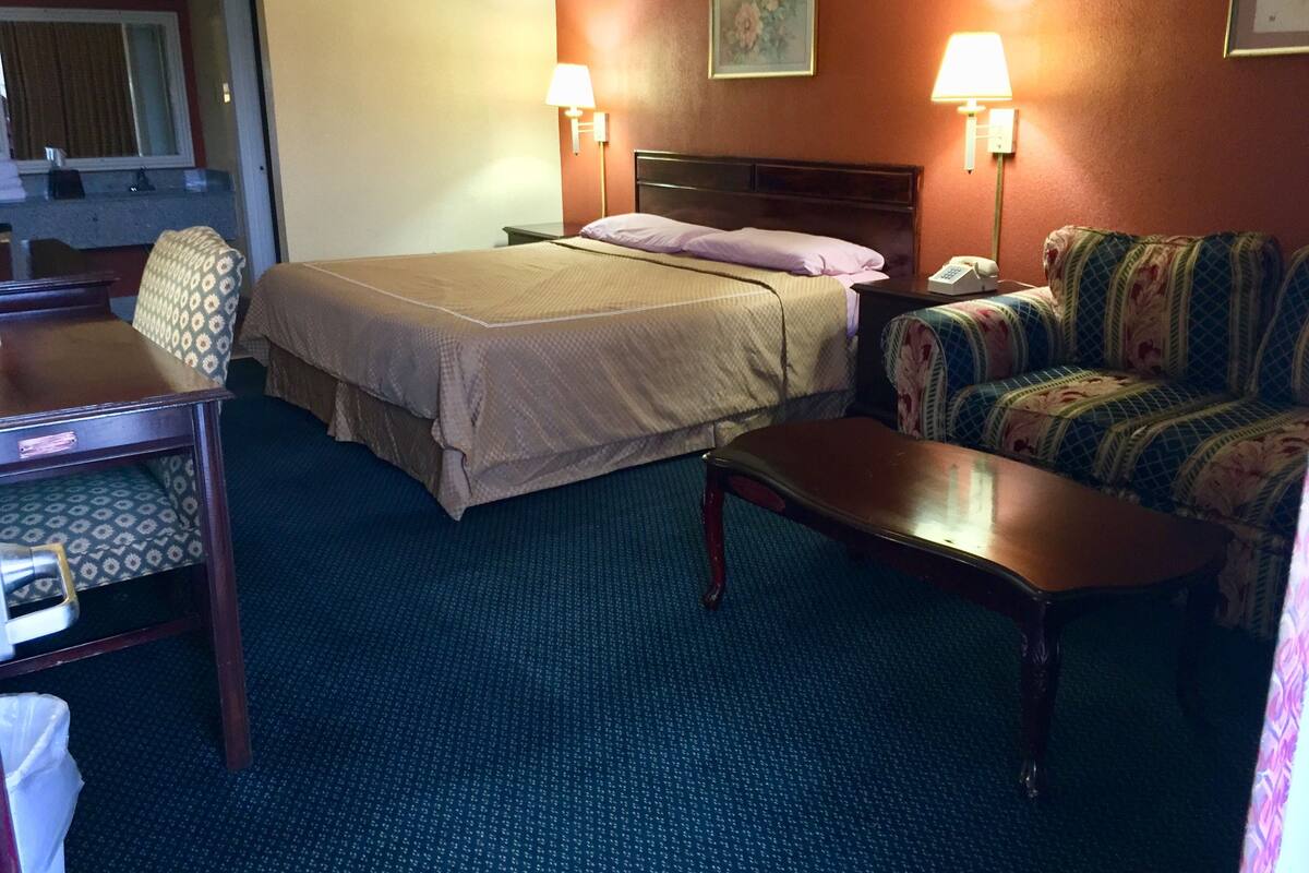 A hotel room with a bed, a desk, a chair, and a sofa. There are two lamps on either side of the bed.