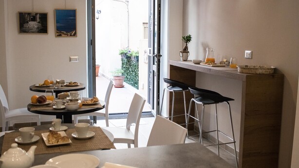 Uma área de refeições com uma mesa posta para o café da manhã, incluindo pratos, xícaras e um bule. Há cadeiras ao redor da mesa e um balcão com banquetas.