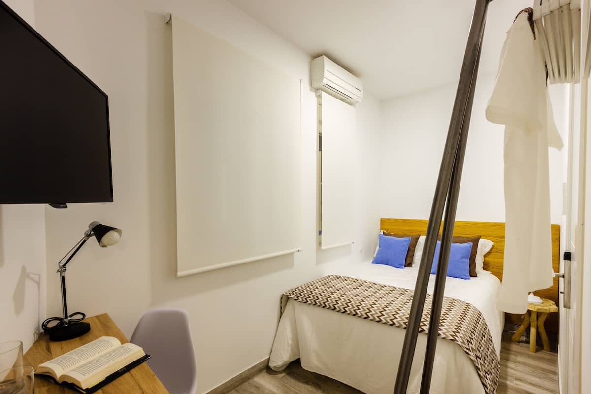 A hotel room with a bed, a desk with a lamp, a book, and an air conditioning unit.