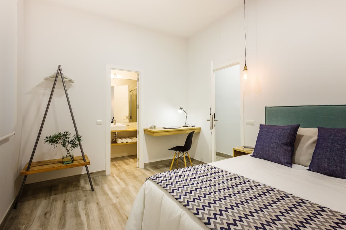 A bedroom with a bed, a desk, a chair, and a hanging plant.