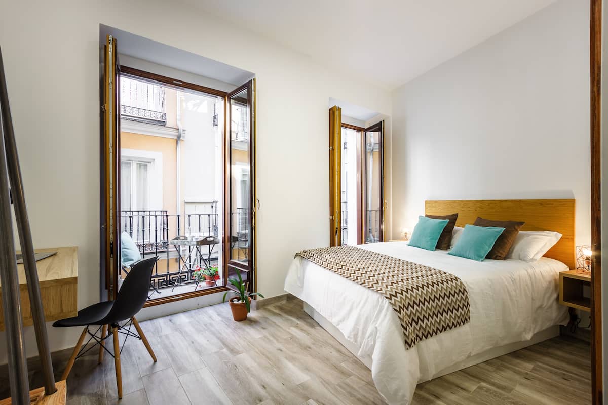A bedroom with a bed, a chair, a small table, and a window with a view of a balcony.