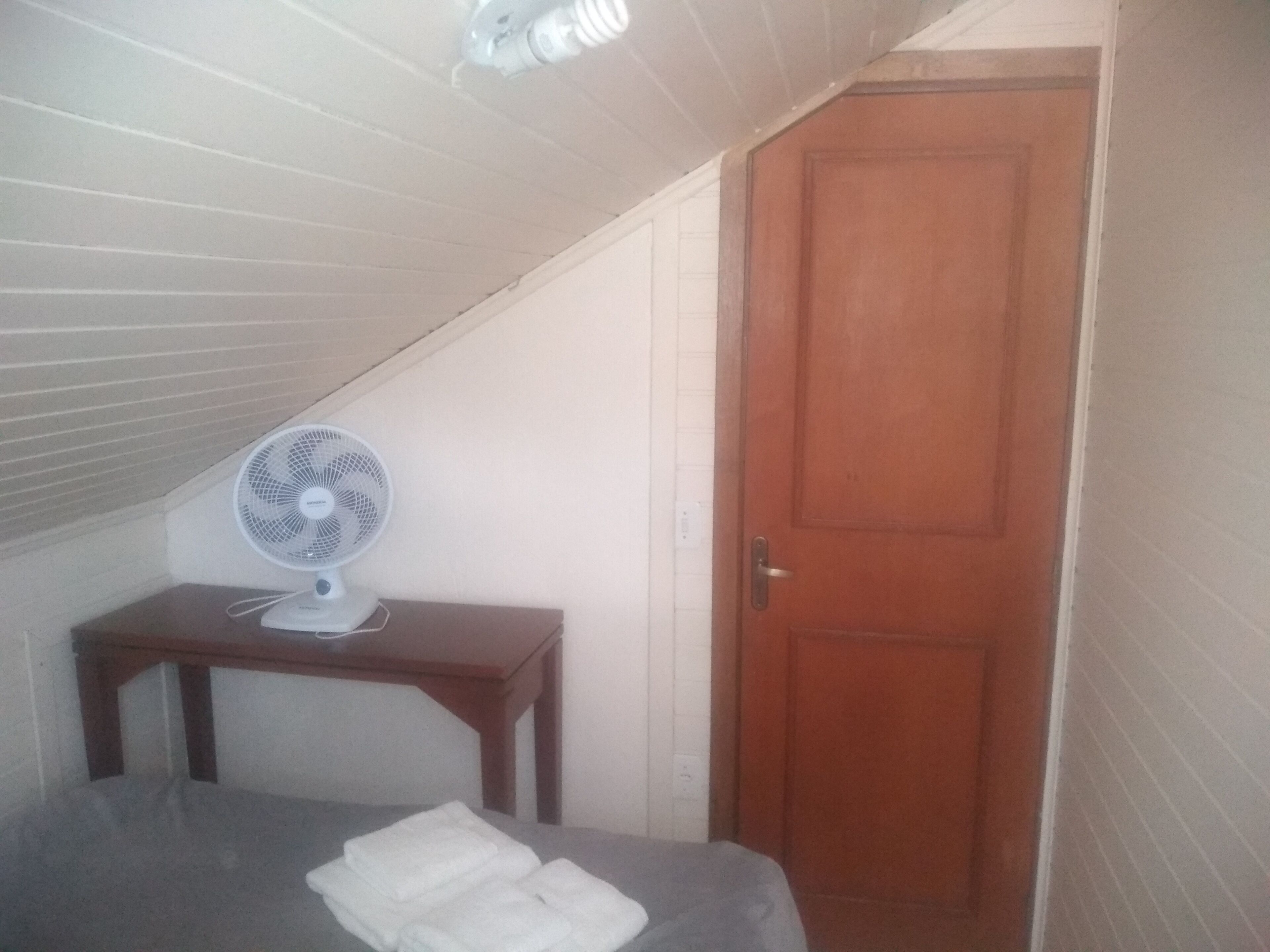 A bedroom with a bed, a wooden door, a small table with a fan, and a sloped ceiling.