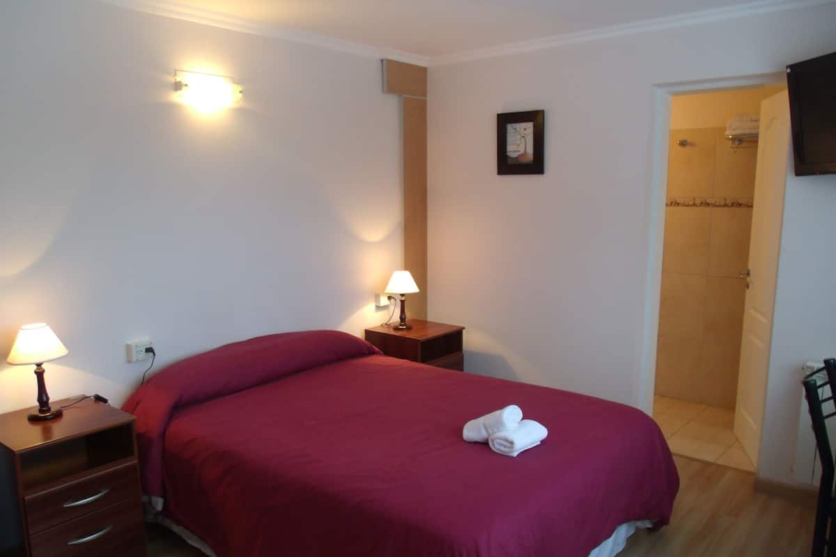 A hotel room with a single bed, two bedside tables with lamps, a small bedside table with a lamp, a framed picture on the wall, and a bathroom door visible in the background.