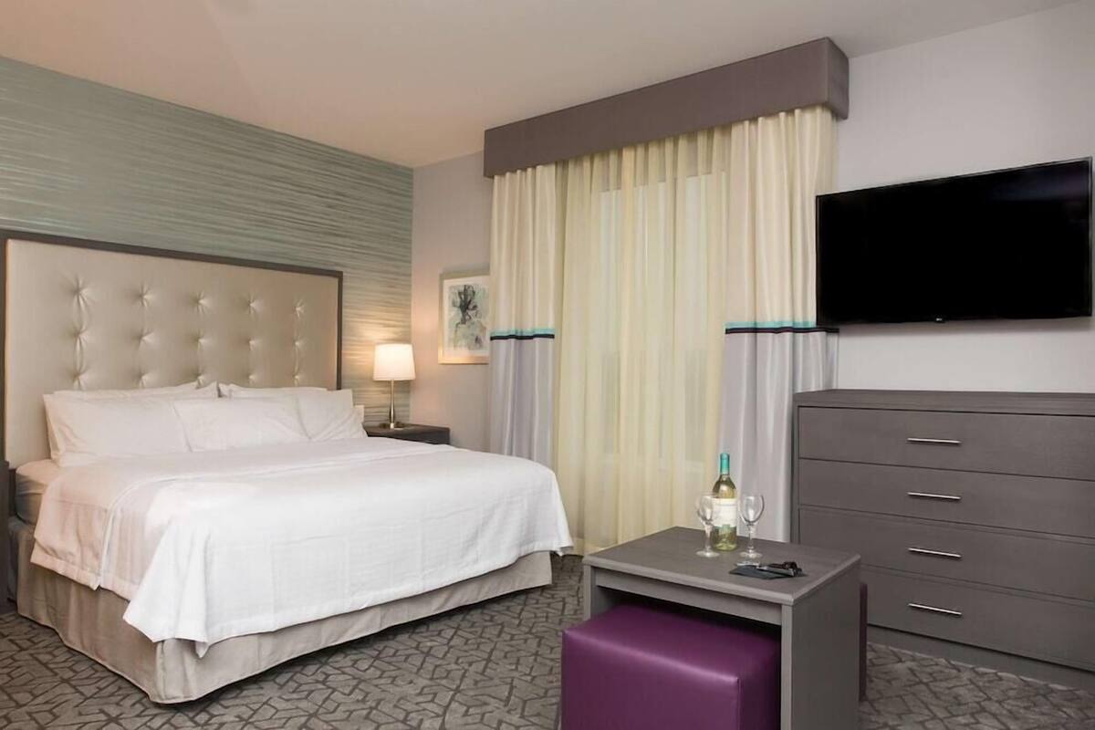 A hotel room with a bed, a desk with a bottle and glass, a TV, and a dresser.