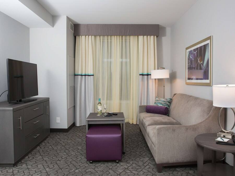 A hotel room with a television on a cabinet, a small table with a bottle and glasses, a sofa with a lamp, and a framed picture on the wall.