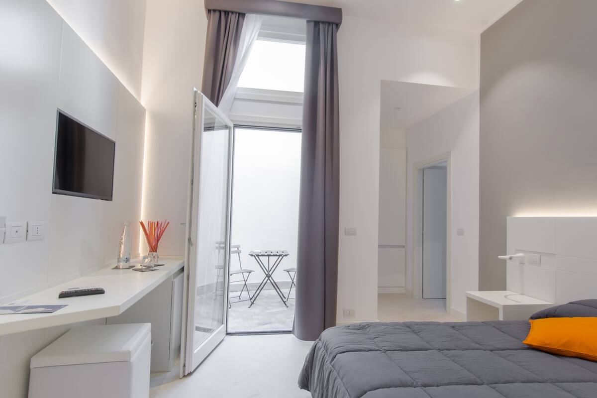 A modern hotel room with a bed, a desk, a TV, and a balcony with a table and chairs.
