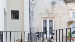 An outdoor patio with a wooden table and chairs, a metal fence, and a building with a window and a door in the background. - Ortigia Bed and Breakfast (Syracuse)