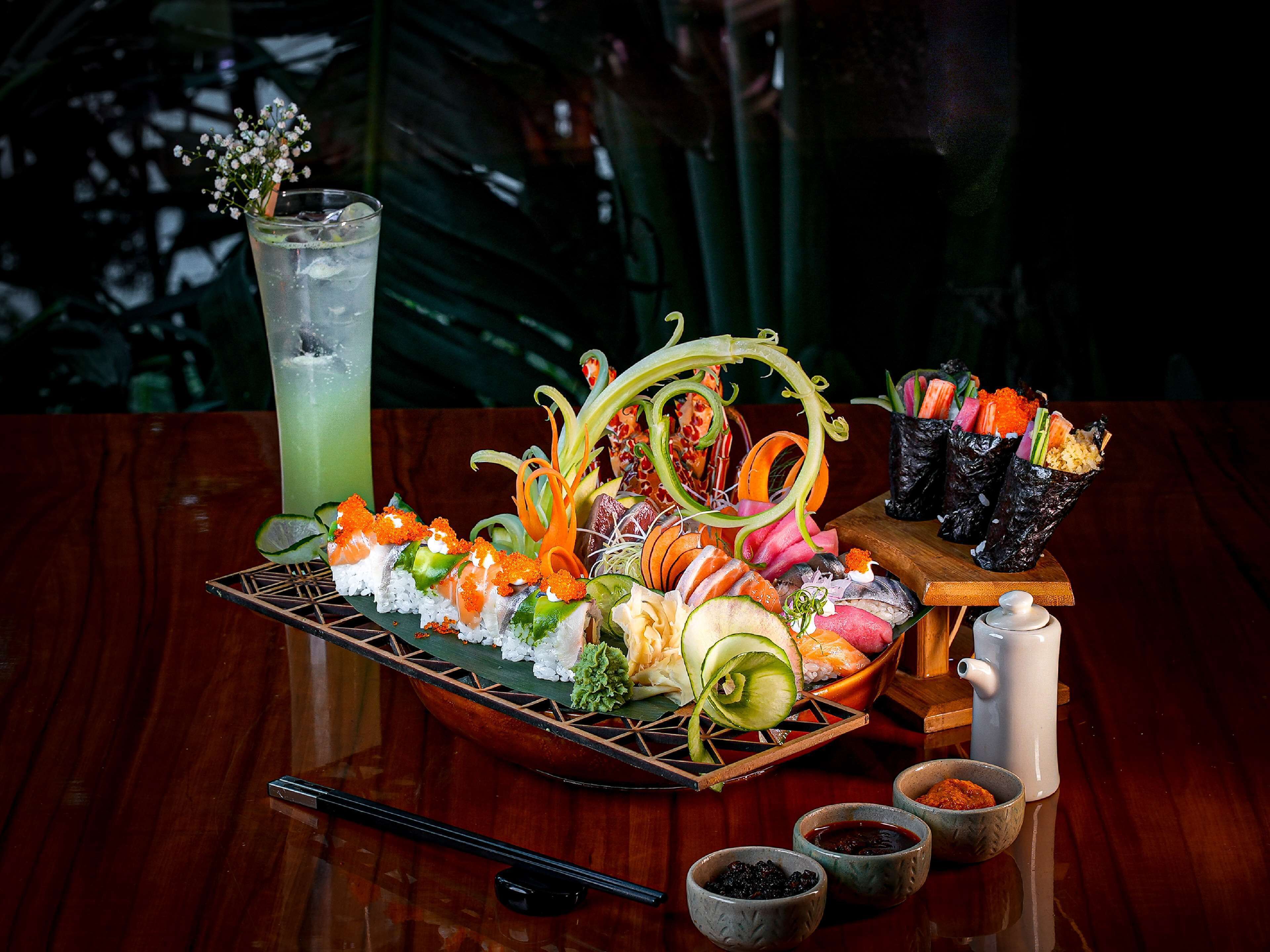 A sushi platter with various types of sushi, a glass of green drink, and small bowls of condiments on a wooden table.