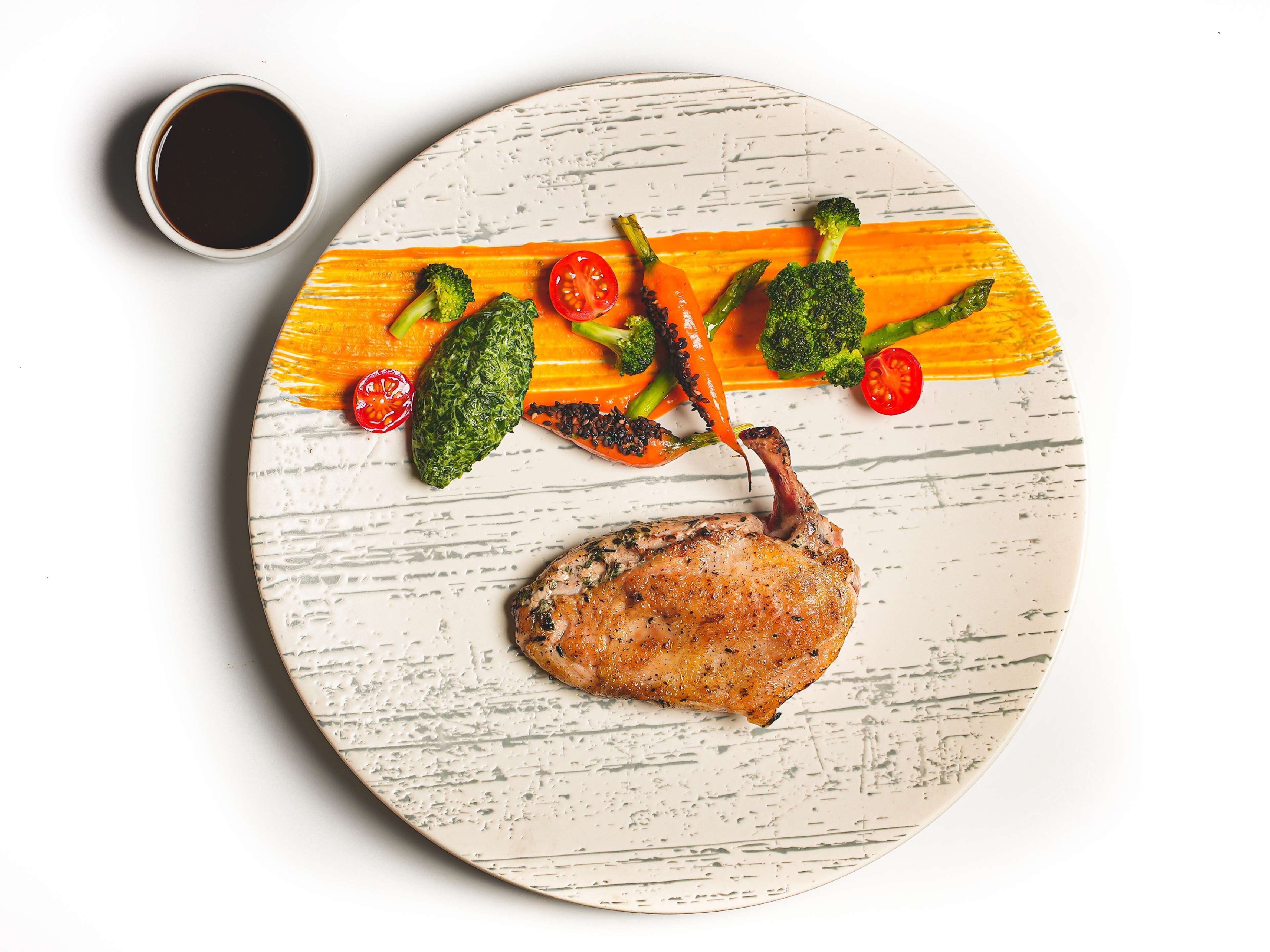 A plate with a roasted duck, broccoli, asparagus, and cherry tomatoes, accompanied by a small cup of sauce.