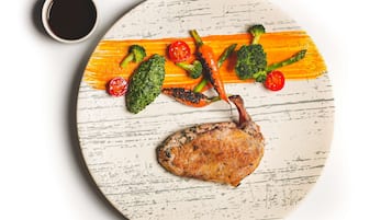 A plate with a roasted duck, broccoli, asparagus, and cherry tomatoes, accompanied by a small cup of sauce.