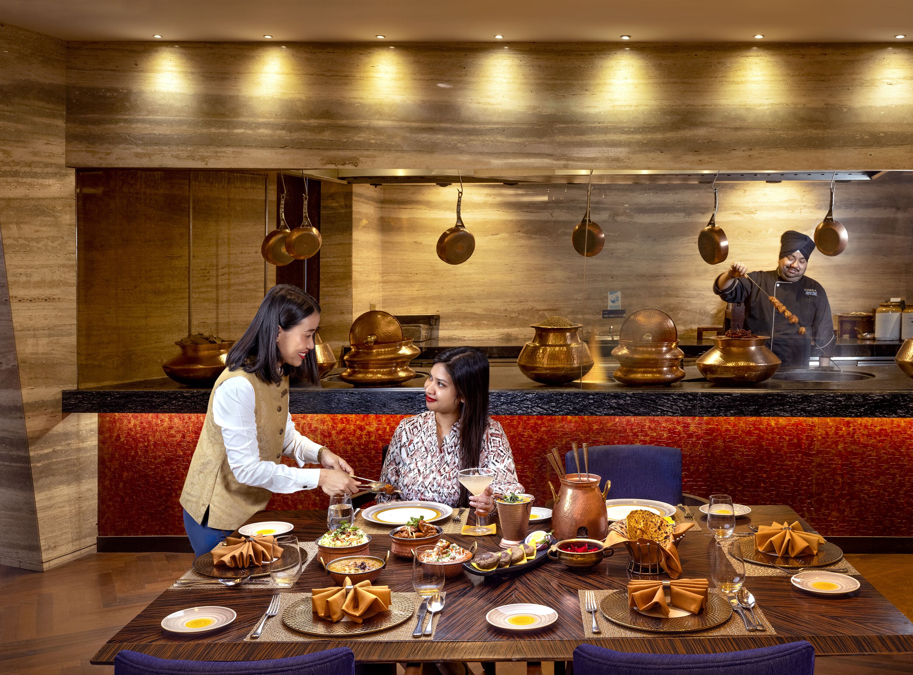Un restaurante con una decoración moderna, una mesa de comedor dispuesta con diversos platos y bebidas, y un cocinero preparando comida al fondo.