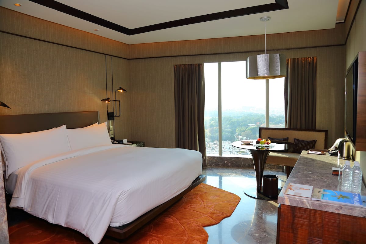 Habitación de hotel con una cama grande, un escritorio con silla, vistas a la ciudad y un ventanal amplio.