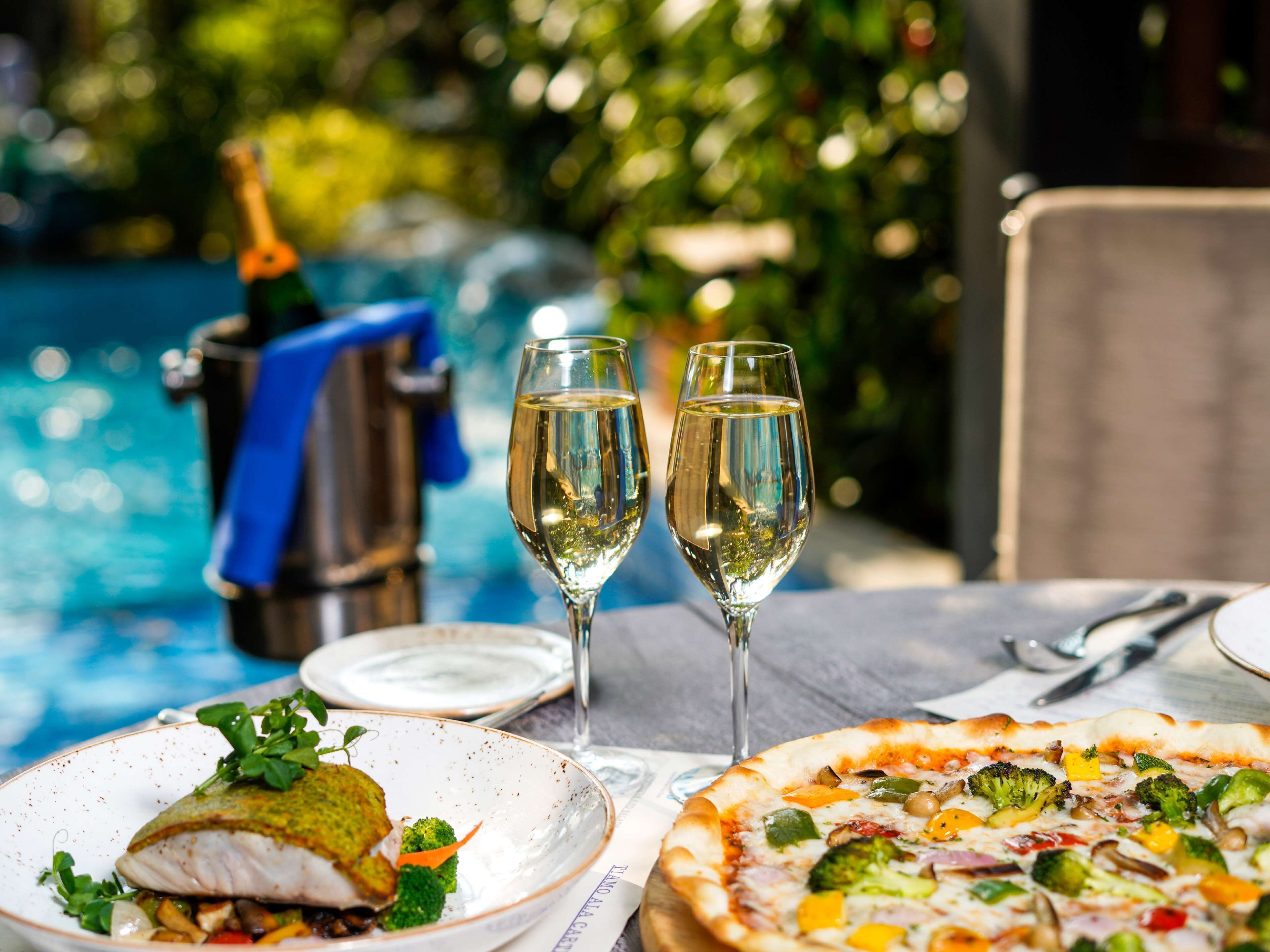 A dining table with a pizza and a fish dish, two glasses of white wine, and a cocktail shaker.