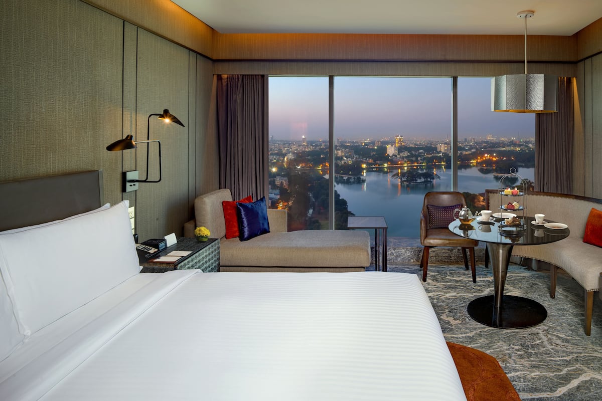 Habitación de hotel con una cama grande, un sofá, una mesita con juego de té y vistas a la ciudad y al agua.