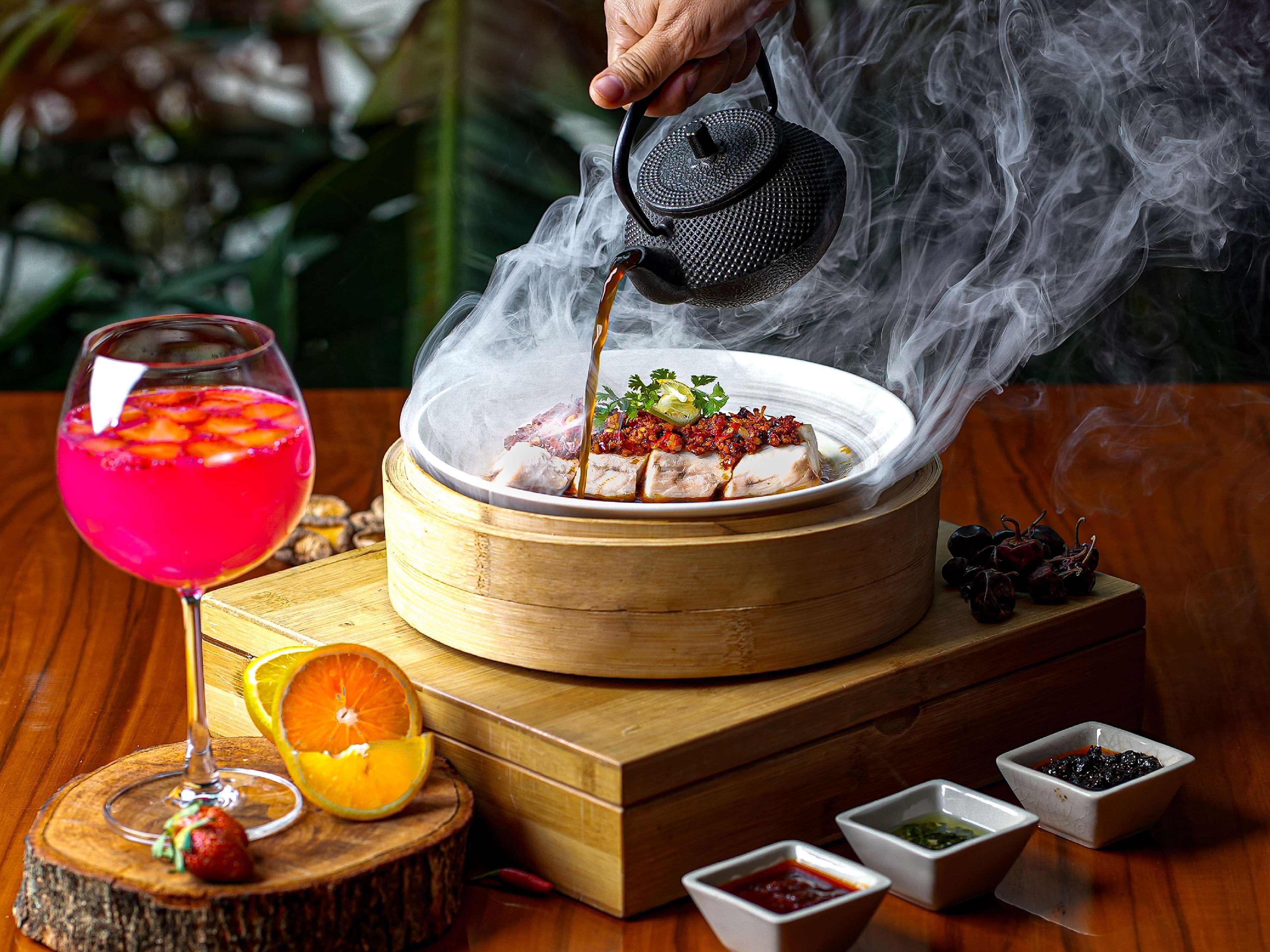 Un plato de olla caliente con vapor saliendo, un vaso de bebida rosada y pequeños recipientes con condimentos sobre una mesa de madera.