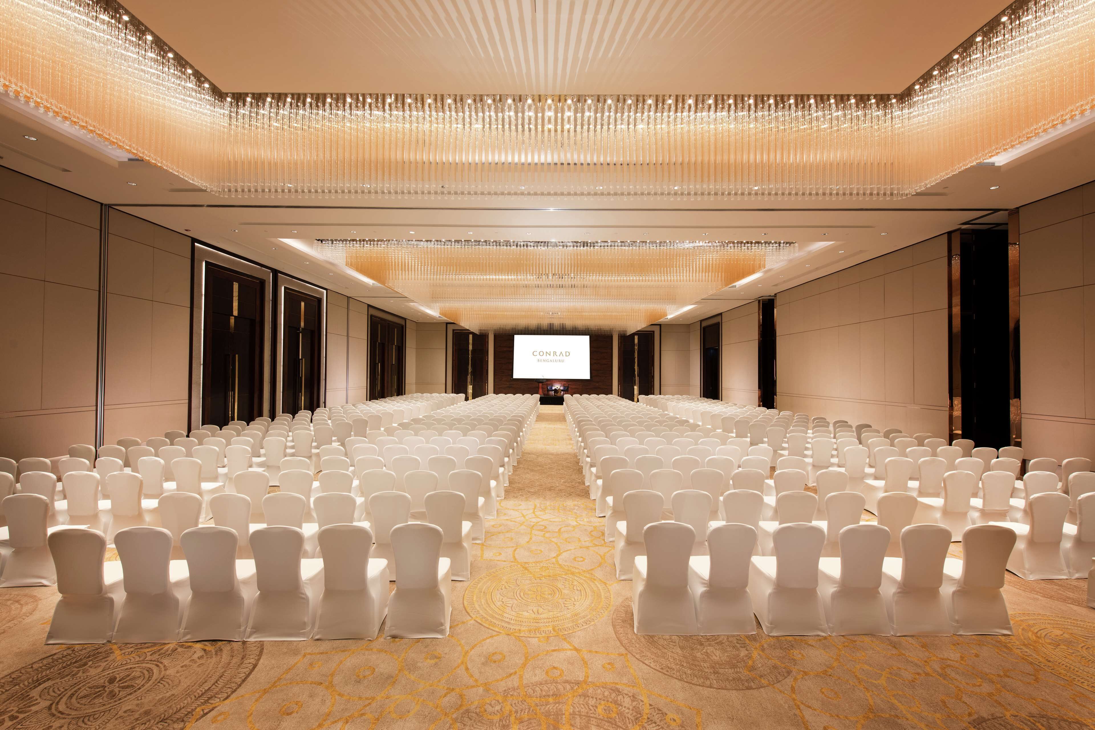 Un salón de conferencias con hileras de sillas blancas, una pantalla grande al frente y una alfombra estampada.