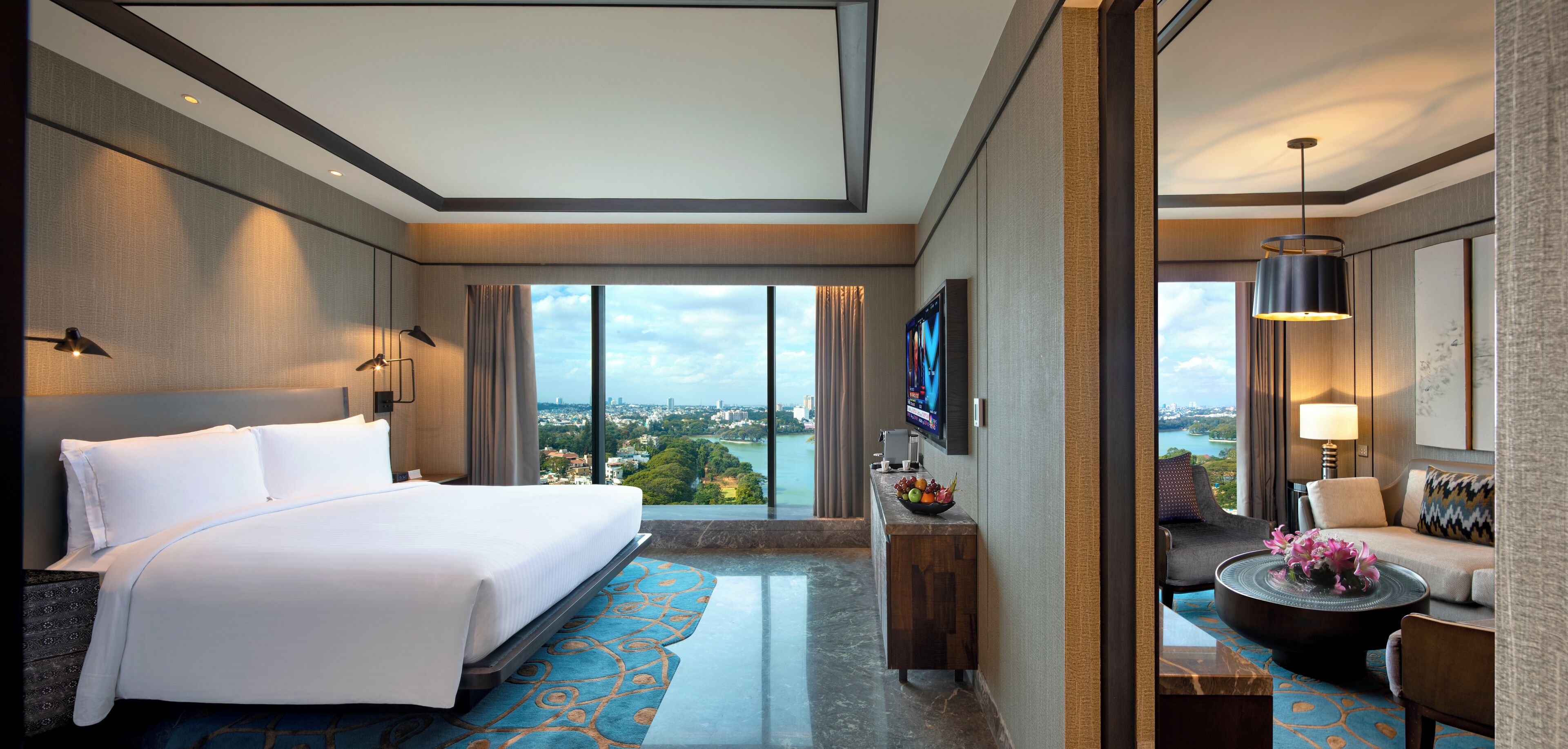 A hotel room with a large bed, a view of the city, a desk with a computer, and a seating area with a lamp.