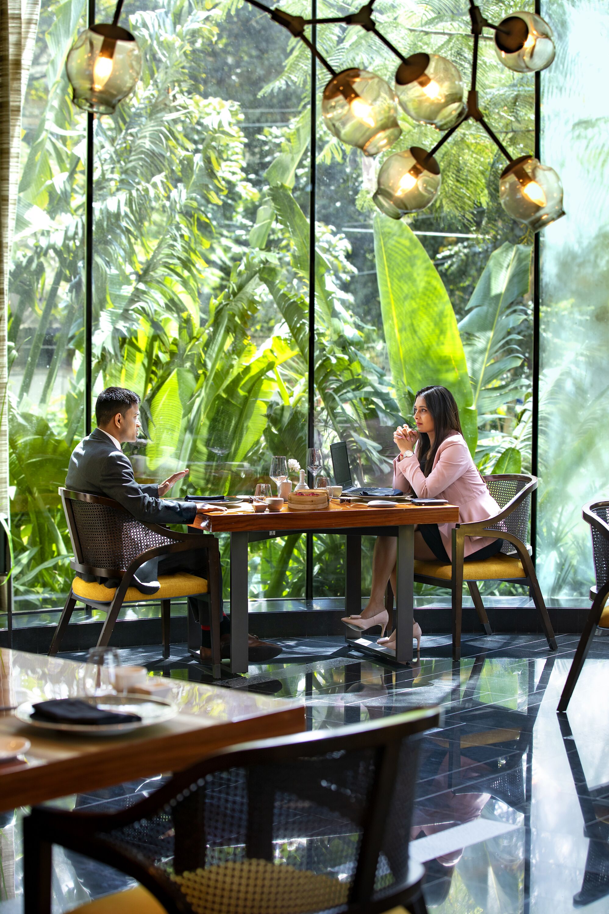 A man and a woman seated at a table in a room with large windows, a glass table, and hanging light fixtures.