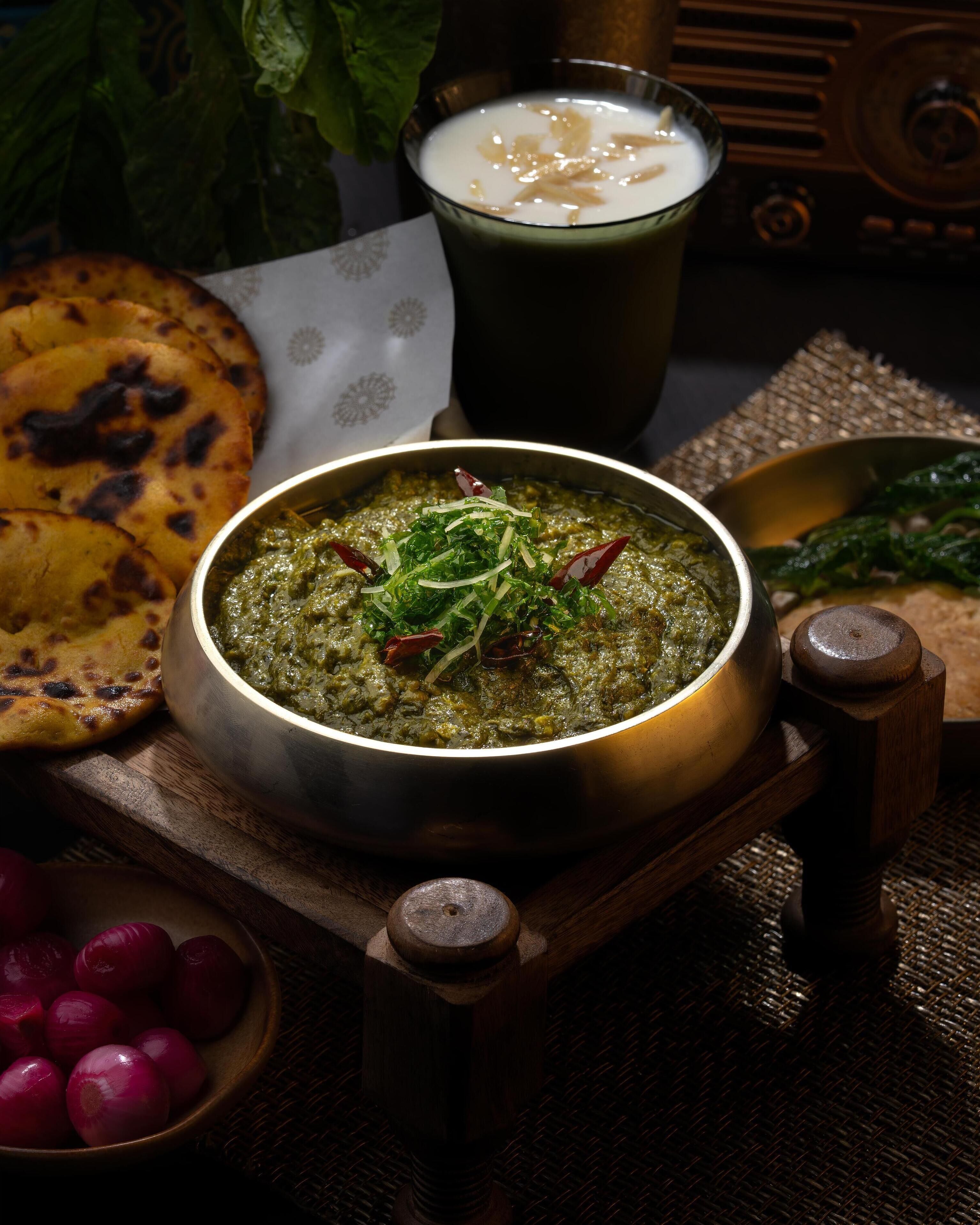 Un tazón de curry verde adornado con hierbas, un vaso de bebida blanca, pan plano y un tazón de cebollas rojas.