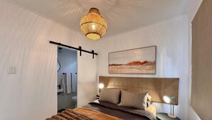 Un dormitorio con una cama, mesitas de noche, una lámpara colgante, un cuadro y una puerta que da al baño.