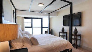 Un dormitorio moderno con una cama grande, una lámpara de noche, un televisor montado en la pared y un ventanal con vistas.