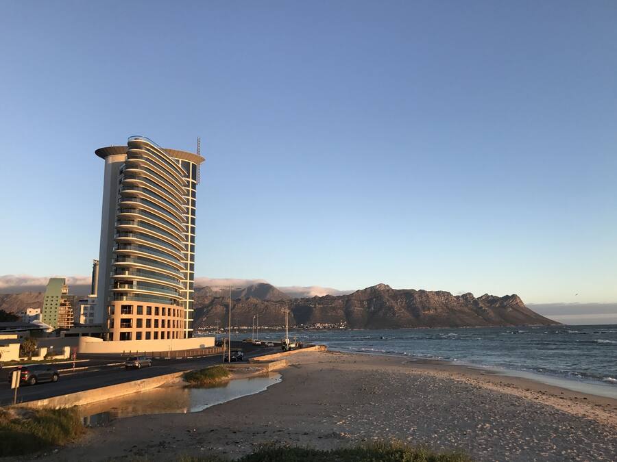 A coastal hotel with a modern design, featuring a curved facade and multiple balconies. The hotel is situated near a beach with a view of the ocean and mountains in the background.