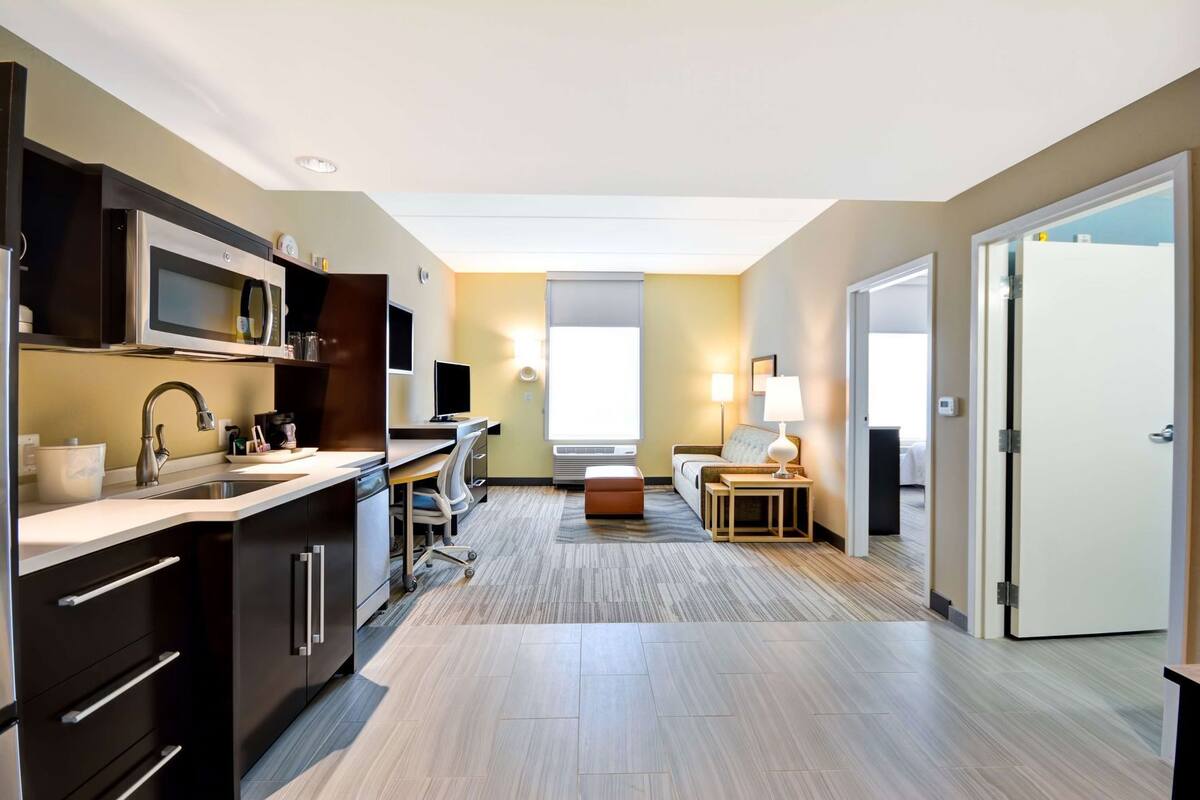 Una habitación de hotel moderna con kitchenette, un escritorio con computadora, una cama y una pequeña zona de estar.