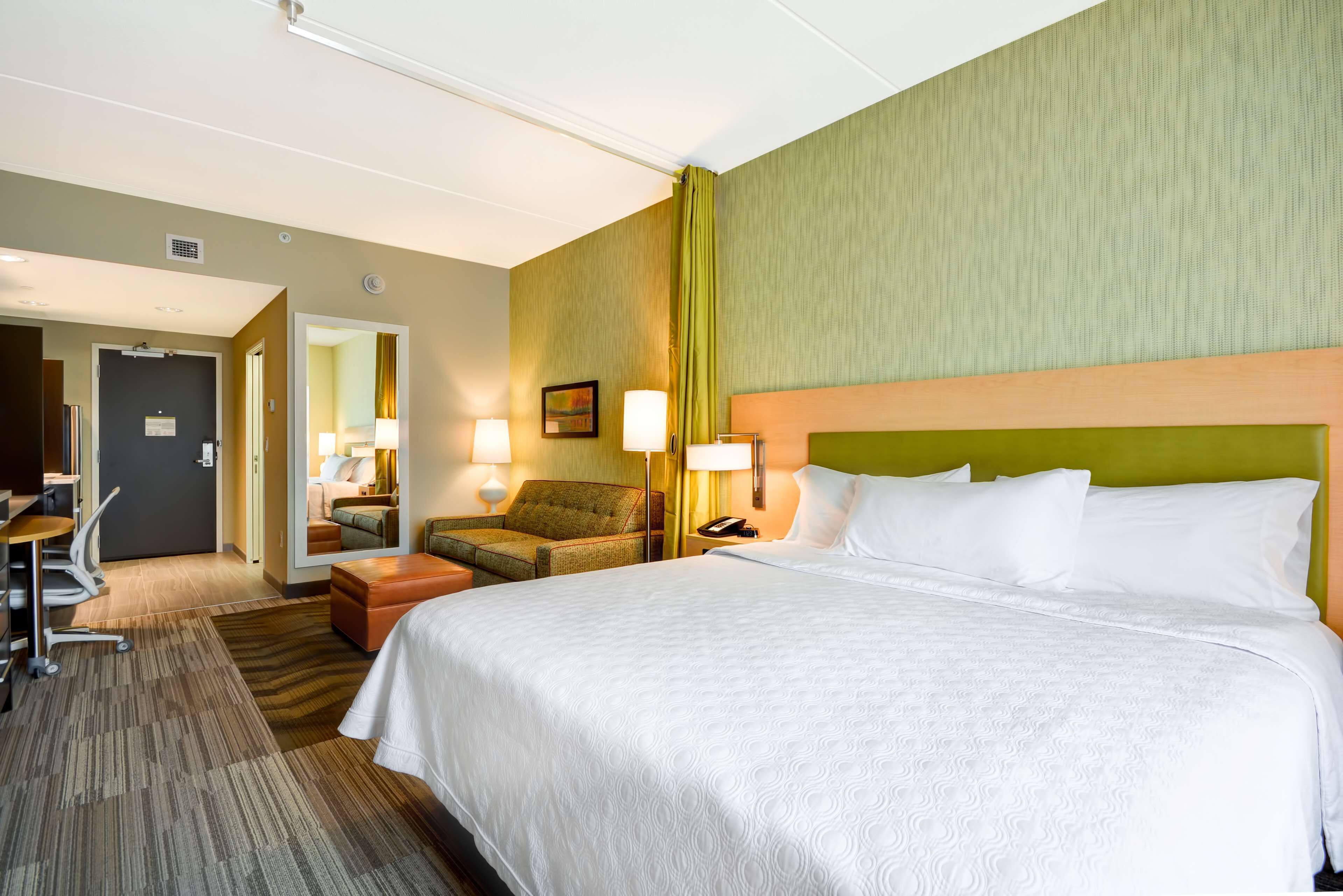 A hotel room with a large bed, a desk with a chair, a small table, and a lamp.