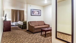 A hotel room with a patterned carpet, a brown sofa, a small table, a framed picture on the wall, and a window with curtains. - Cobblestone Hotel & Suites - Gering/Scottsbluff (Gering)