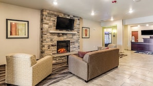 A living room with a stone fireplace, a flat-screen TV, and a reception desk in the background. - Cobblestone Hotel & Suites - Gering/Scottsbluff (Gering)