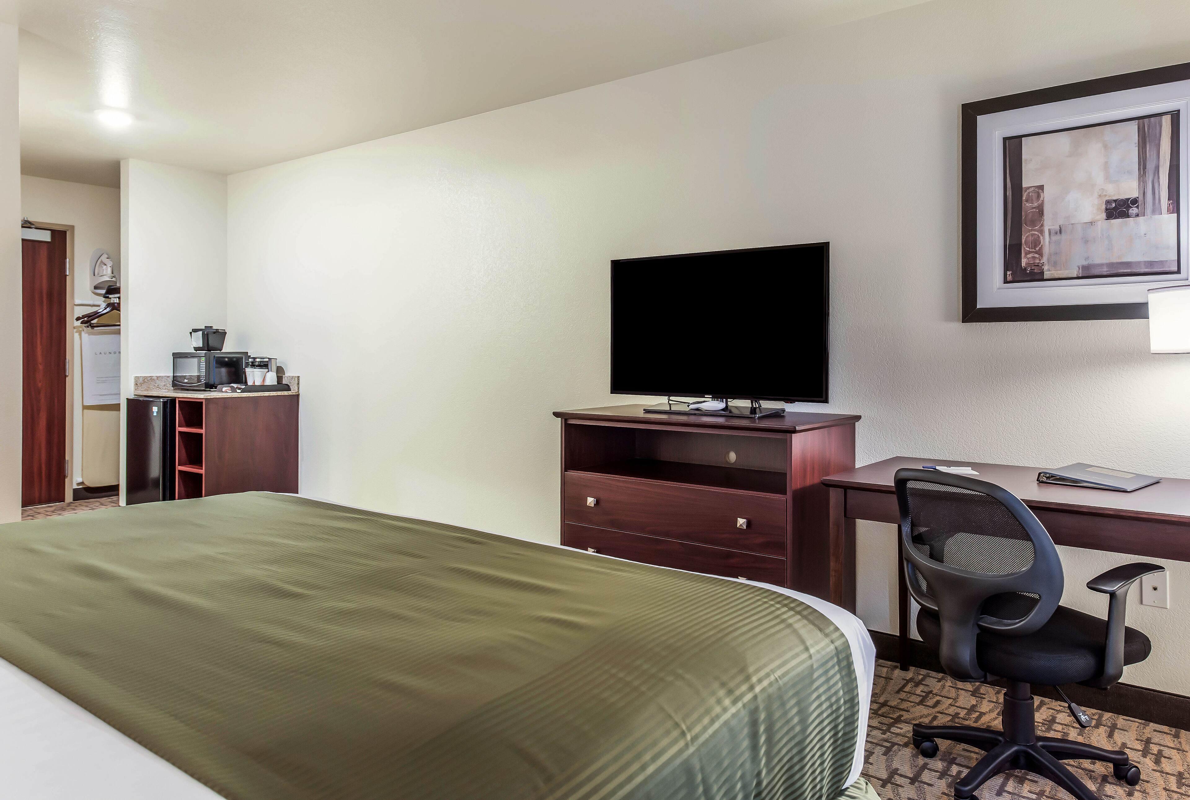A hotel room with a bed, a desk with a computer, a chair, a television, and a small kitchenette.