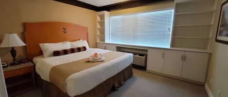 A hotel room with a large bed, a nightstand, a lamp, a window with blinds, and built-in storage cabinets.