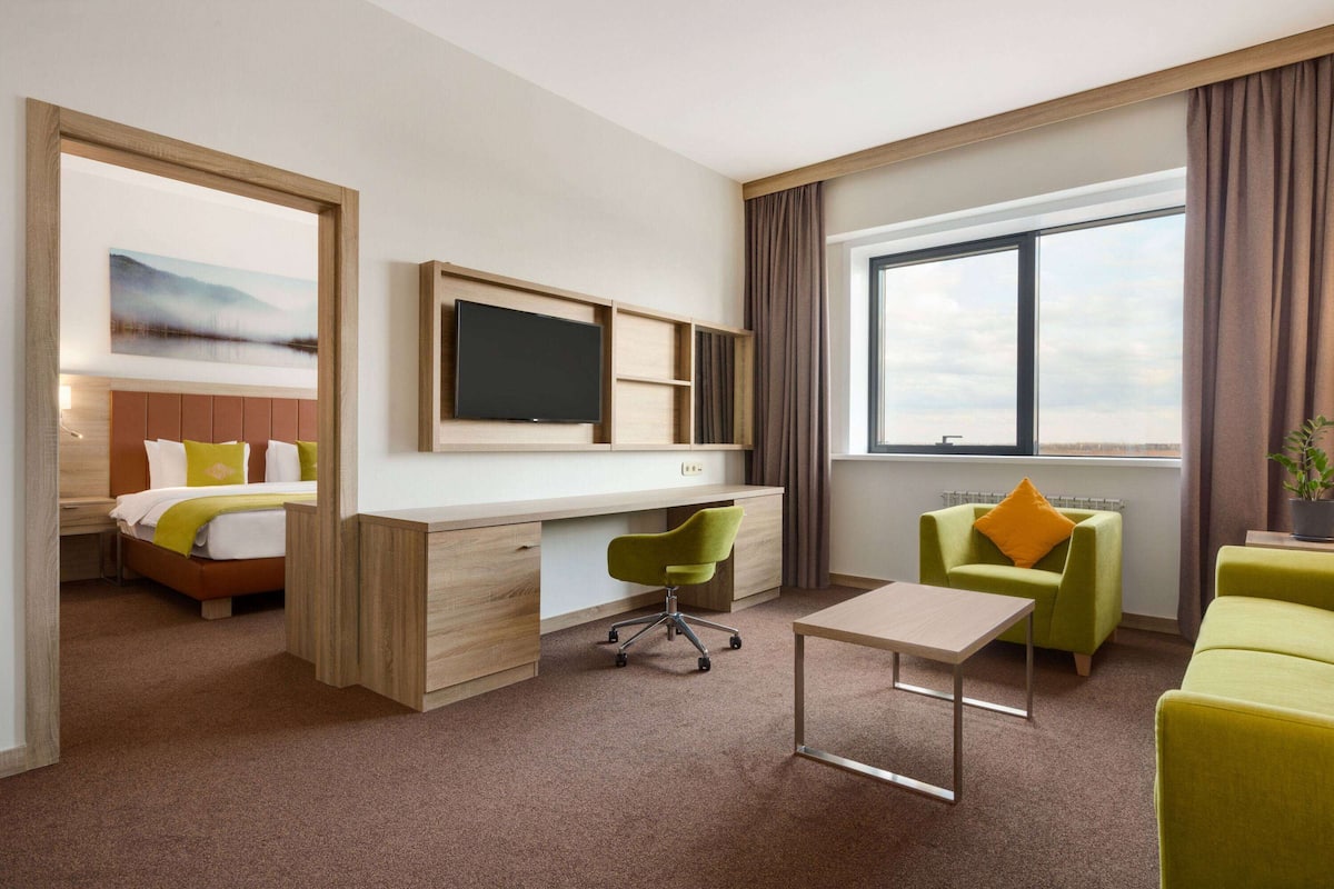 Una habitación de hotel con cama, escritorio, televisor, sillas verdes y un ventanal grande.