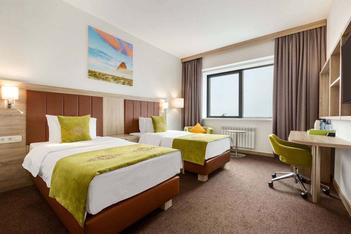 Habitación de hotel con dos camas, un escritorio, una silla, un ventanal y un cuadro en la pared.