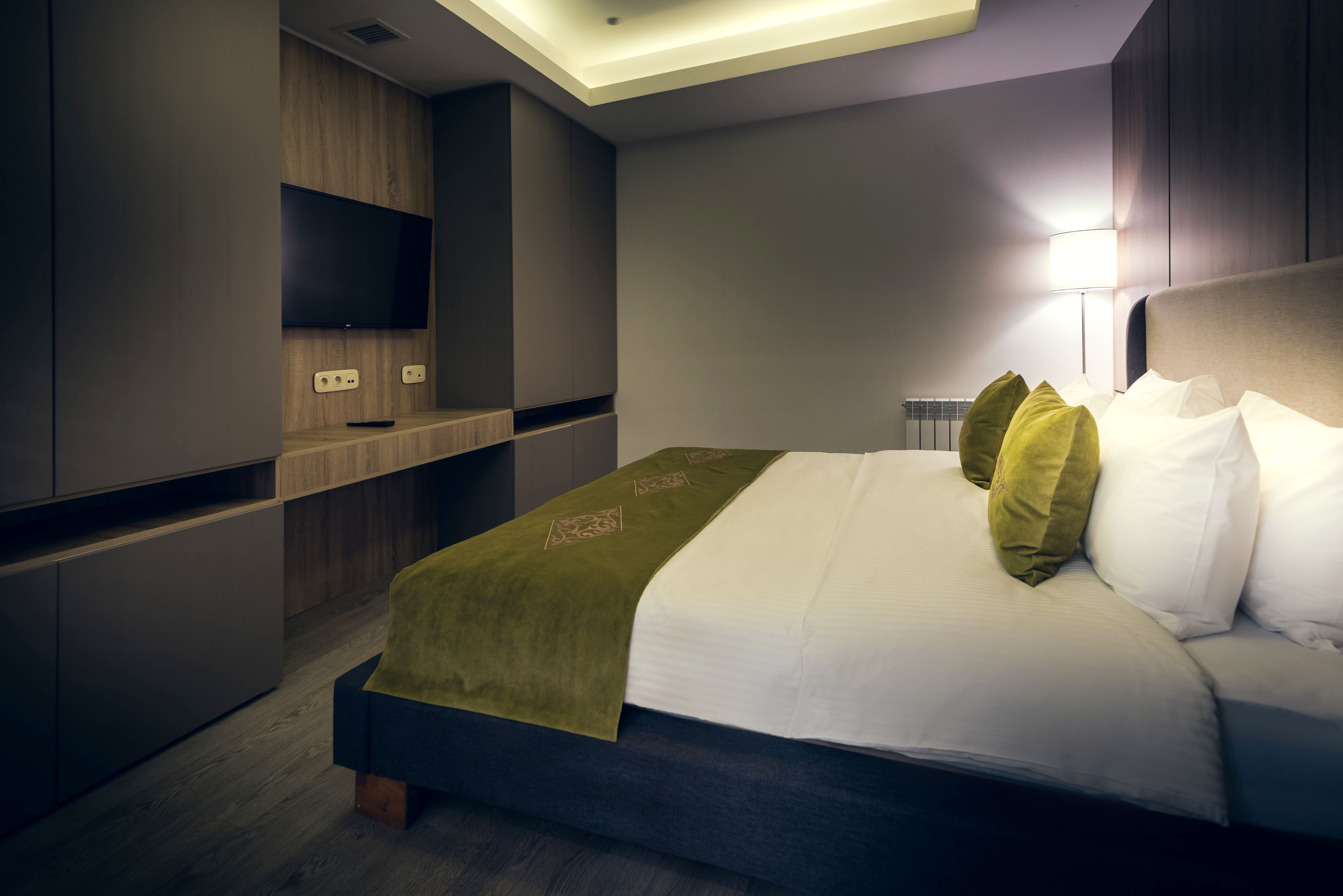 A modern hotel room with a bed, a flat-screen TV mounted on a wooden panel, and a bedside table with a lamp.