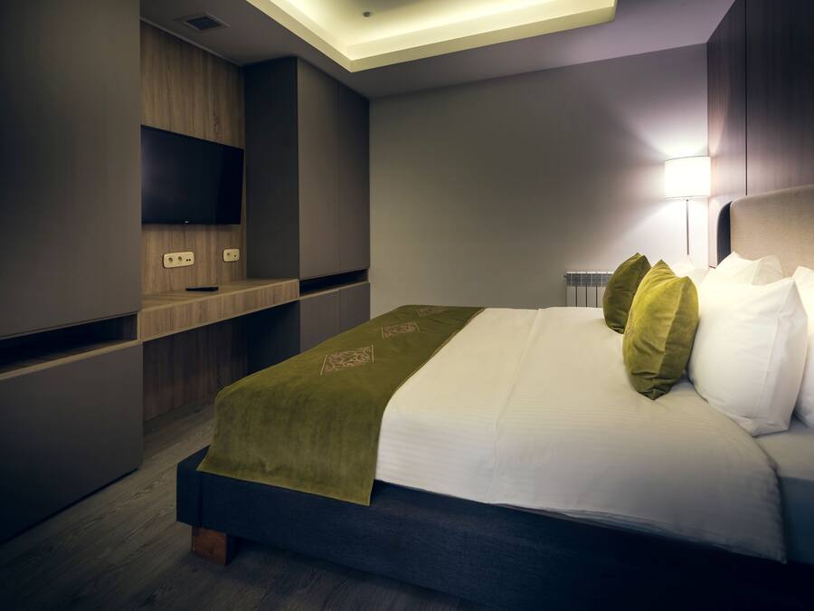 A modern hotel room with a bed, a flat-screen TV mounted on a wooden panel, and a bedside table with a lamp.