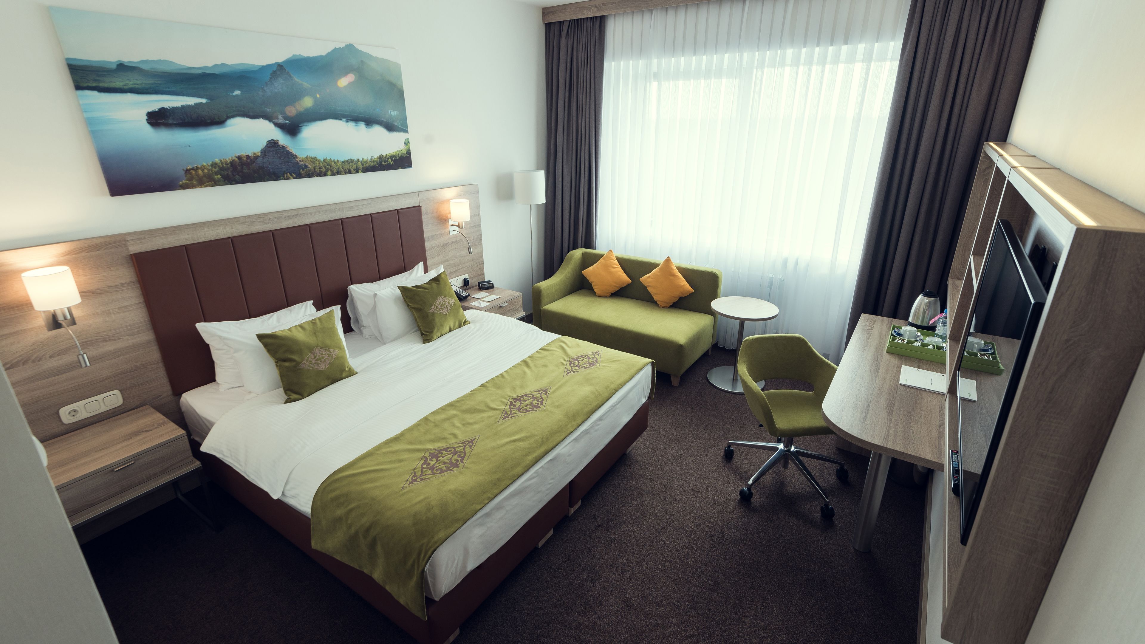 A hotel room with a large bed, a sofa, a desk with a chair, a TV, and a painting on the wall.