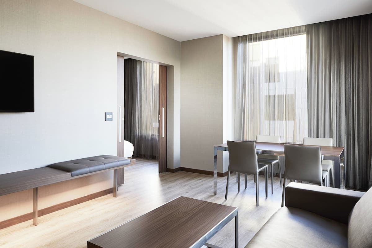 A modern hotel room with a long bench, a small table, a sofa, and a dining area with chairs and a table.
