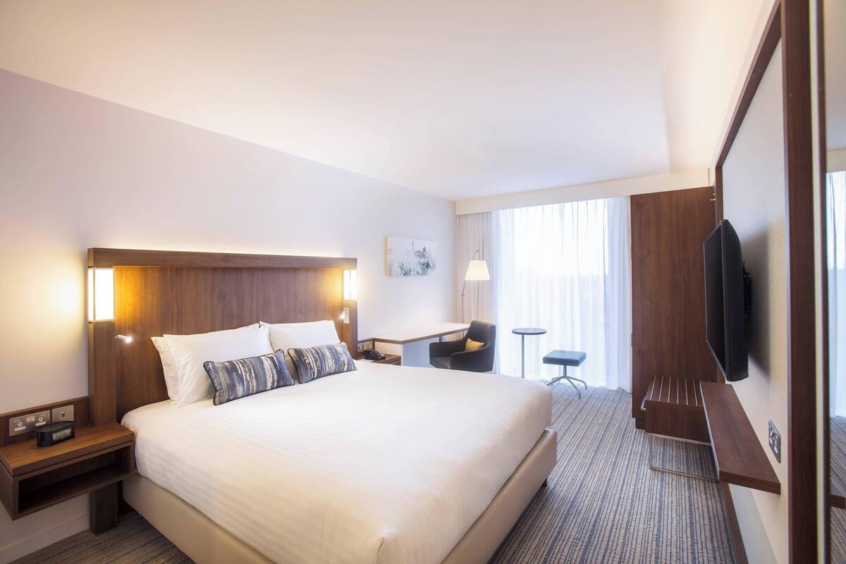A hotel room with a large bed, bedside tables, a desk, a chair, and a TV mounted on the wall.