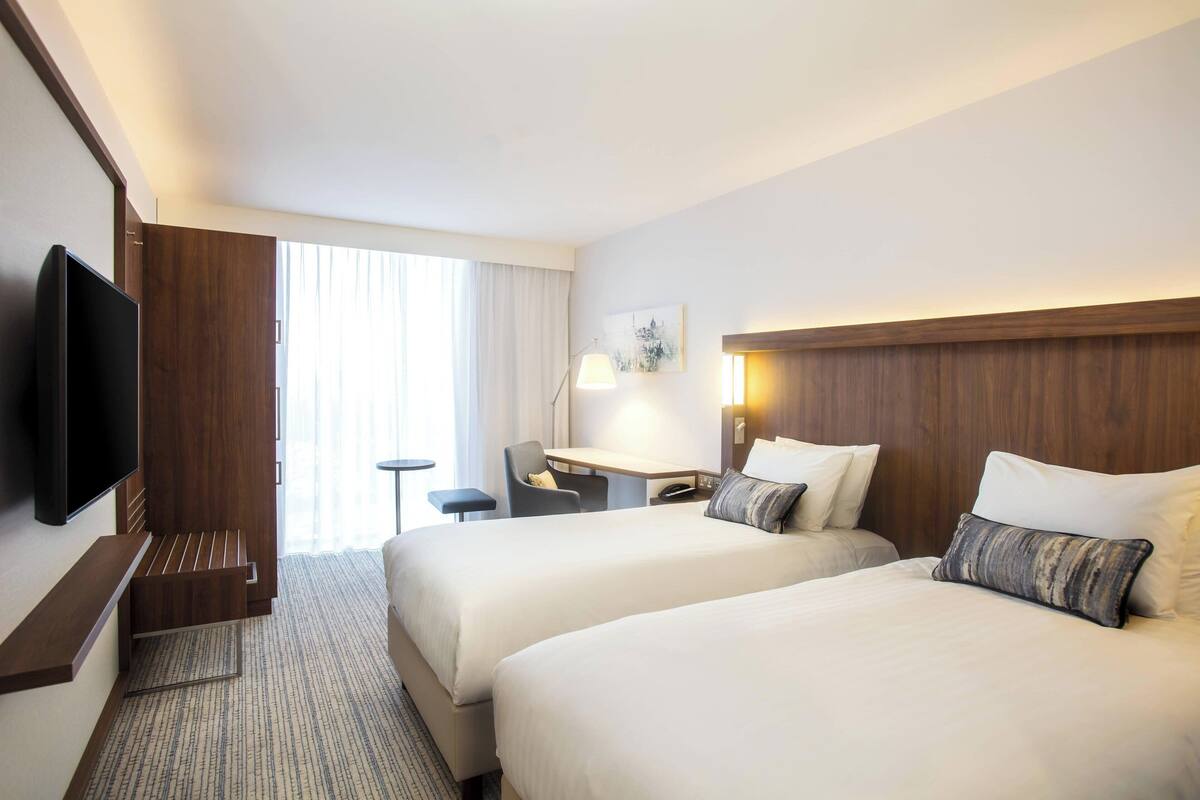 A hotel room with two beds, a desk, a chair, a TV, and a large window with curtains.