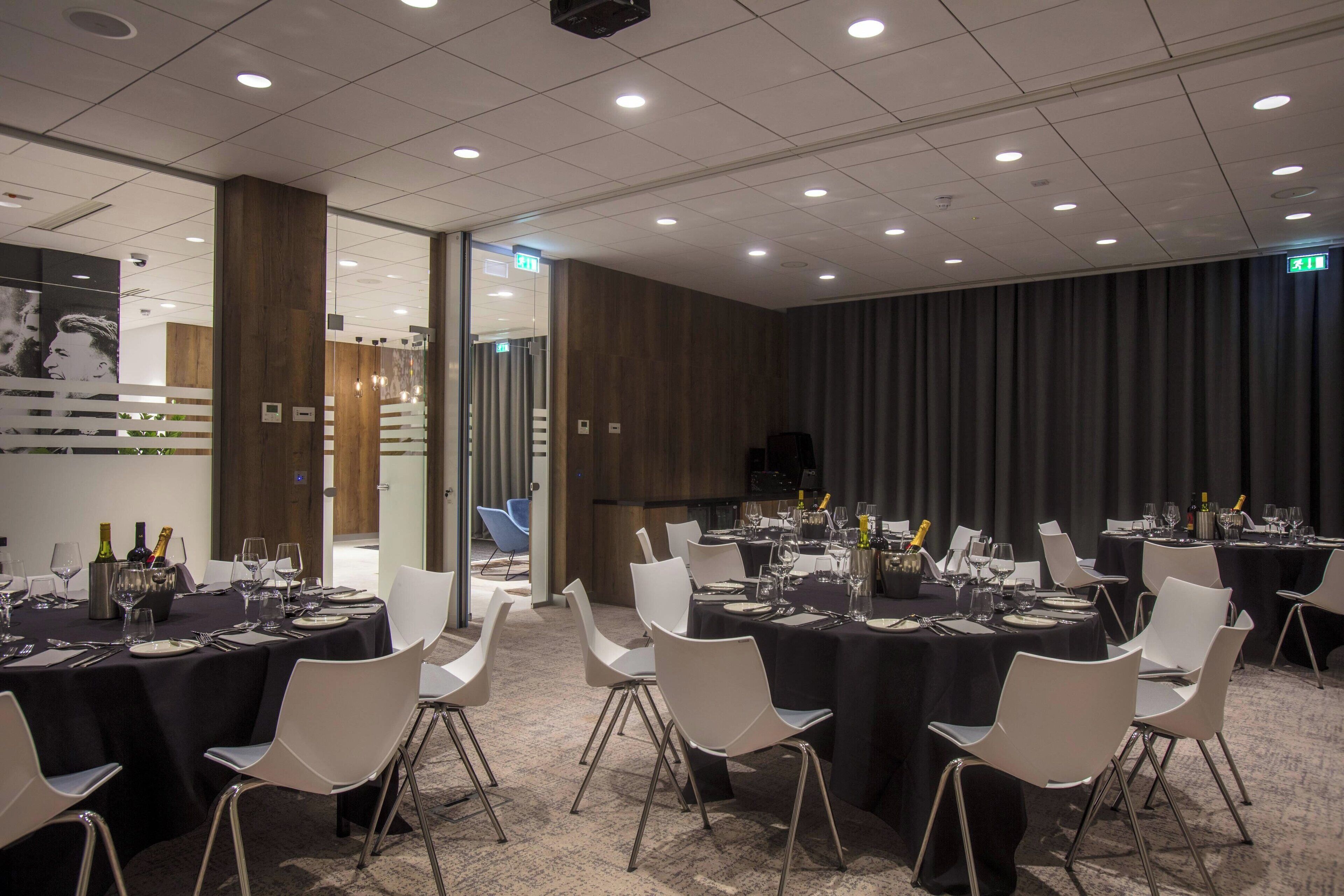 Een congresruimte met meerdere tafels gedekt voor diner, witte stoelen en een donkere gordijnenachtergrond.