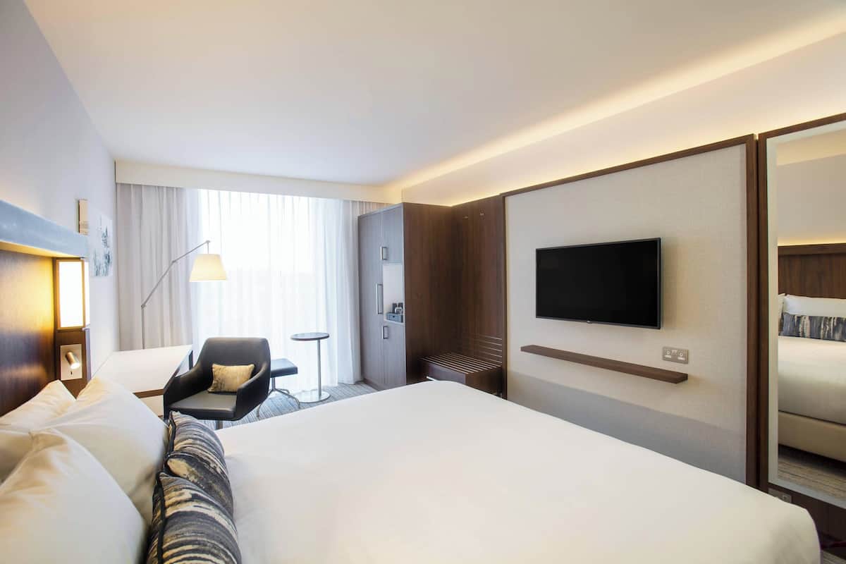 A hotel room with a large bed, a flat-screen TV mounted on the wall, a desk with a chair, and a window with curtains.