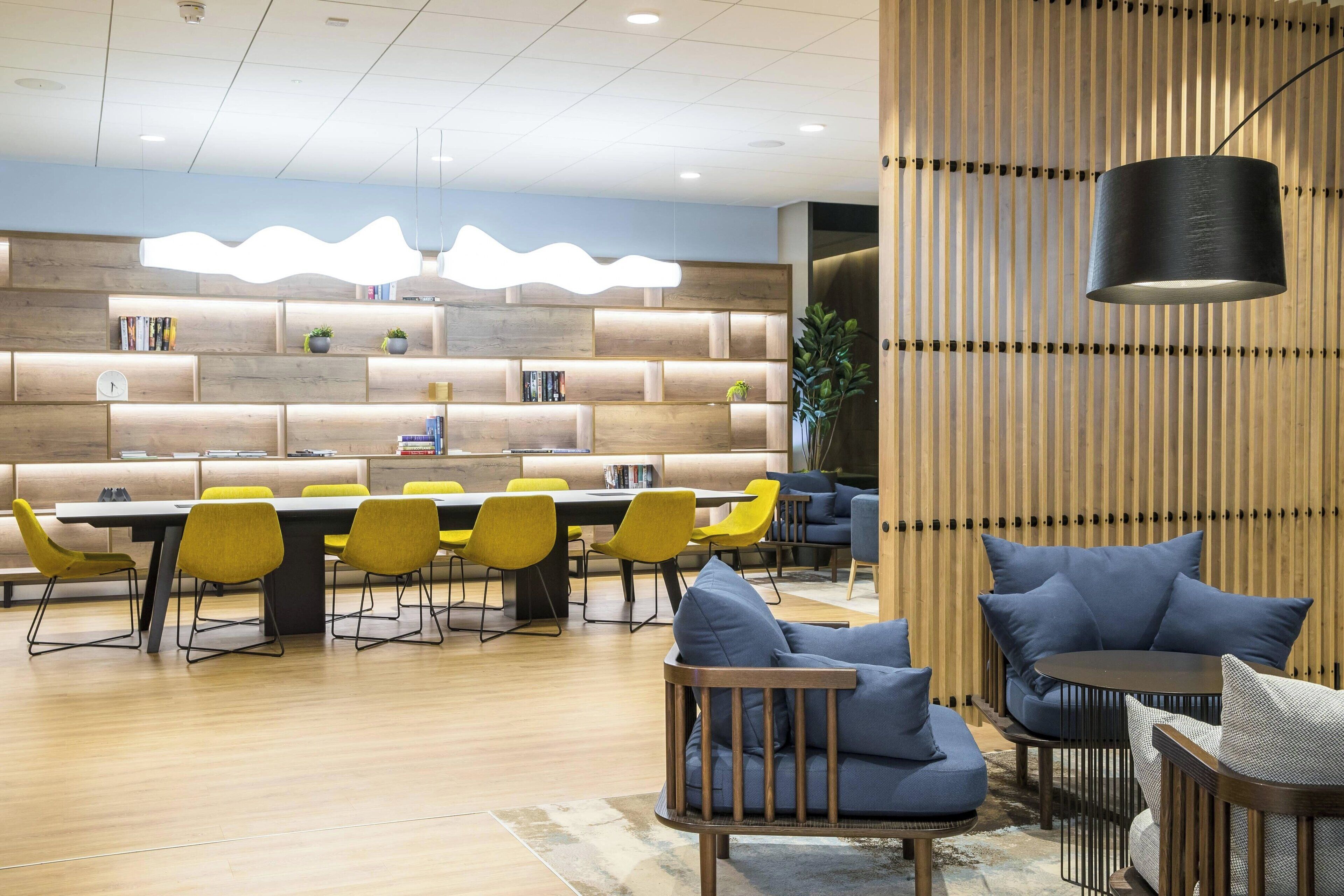 A modern hotel lobby with a wooden reception desk, yellow chairs, a long dining table, and blue sofas.