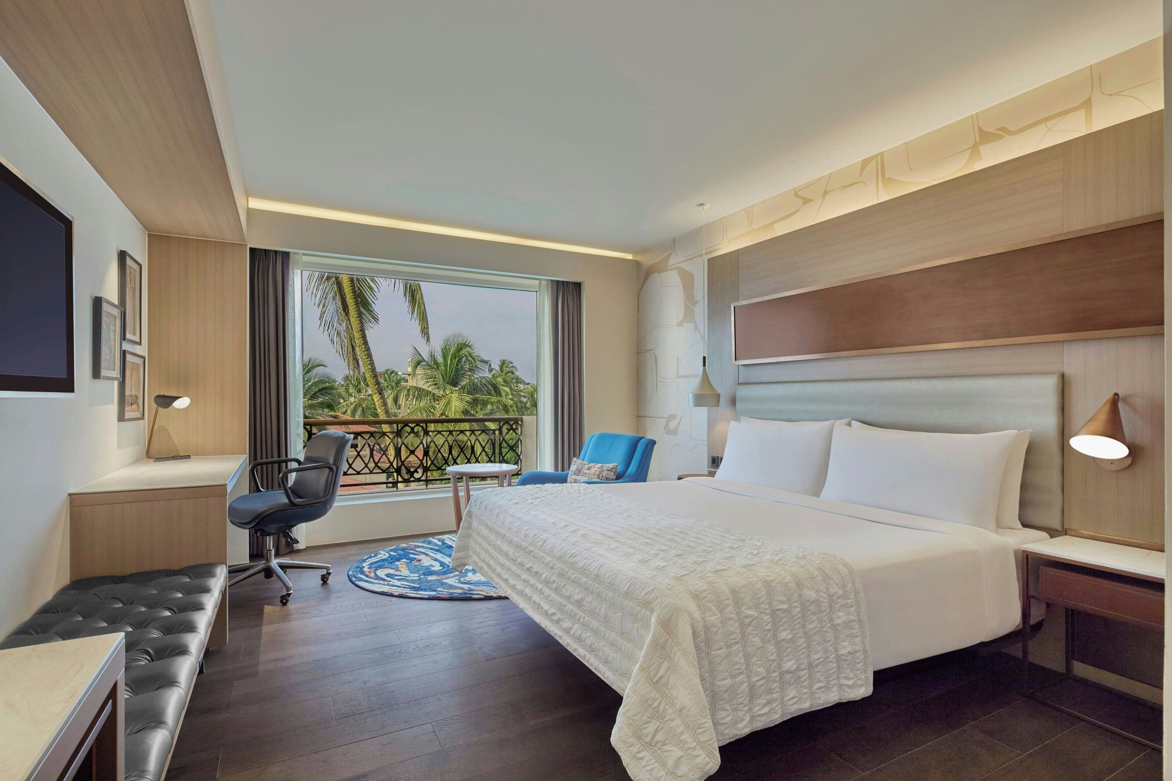 A hotel room with a large bed, a desk, a chair, and a view of palm trees.