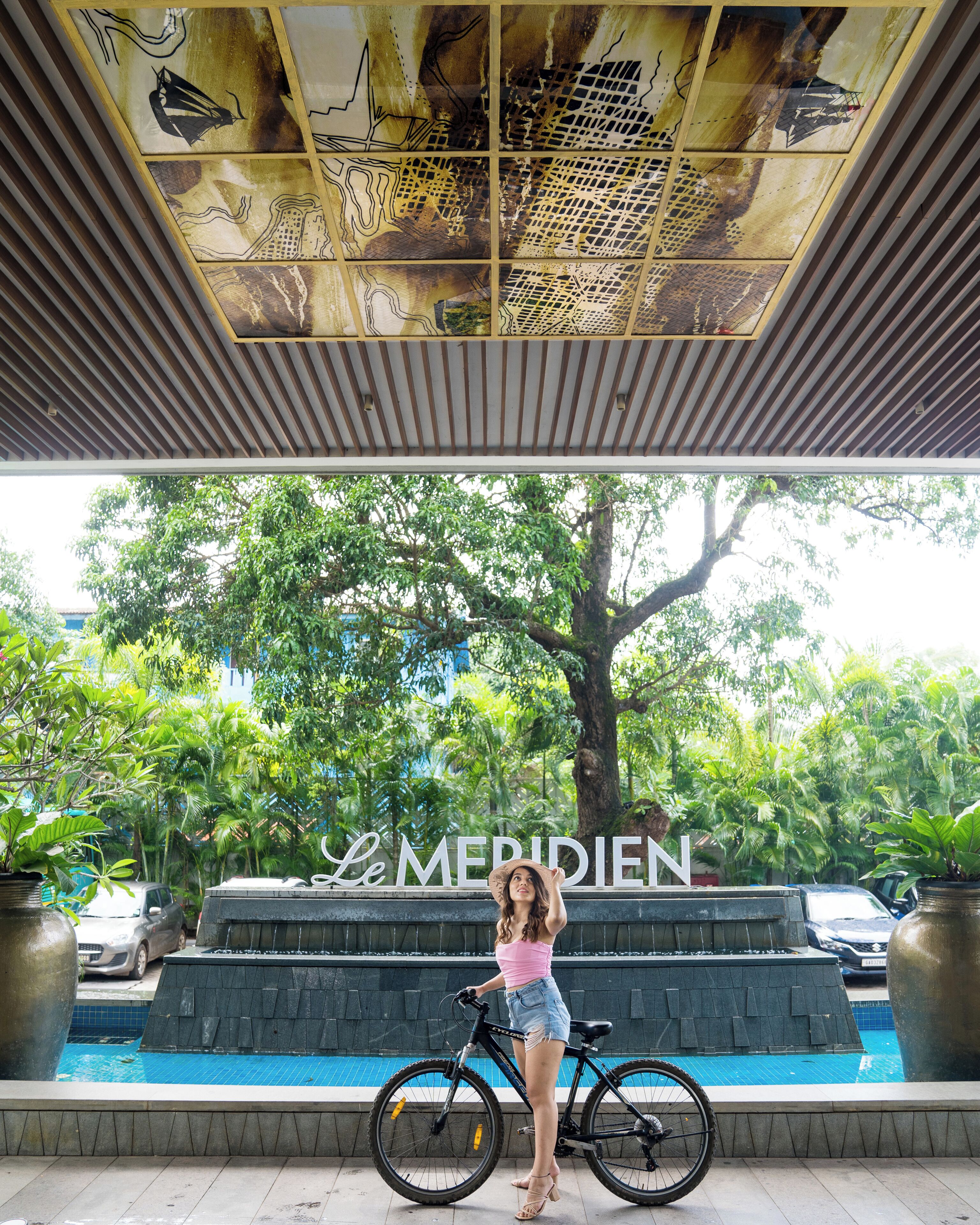 A woman standing next to a bicycle in front of a pool with a sign reading "Le Meridien".