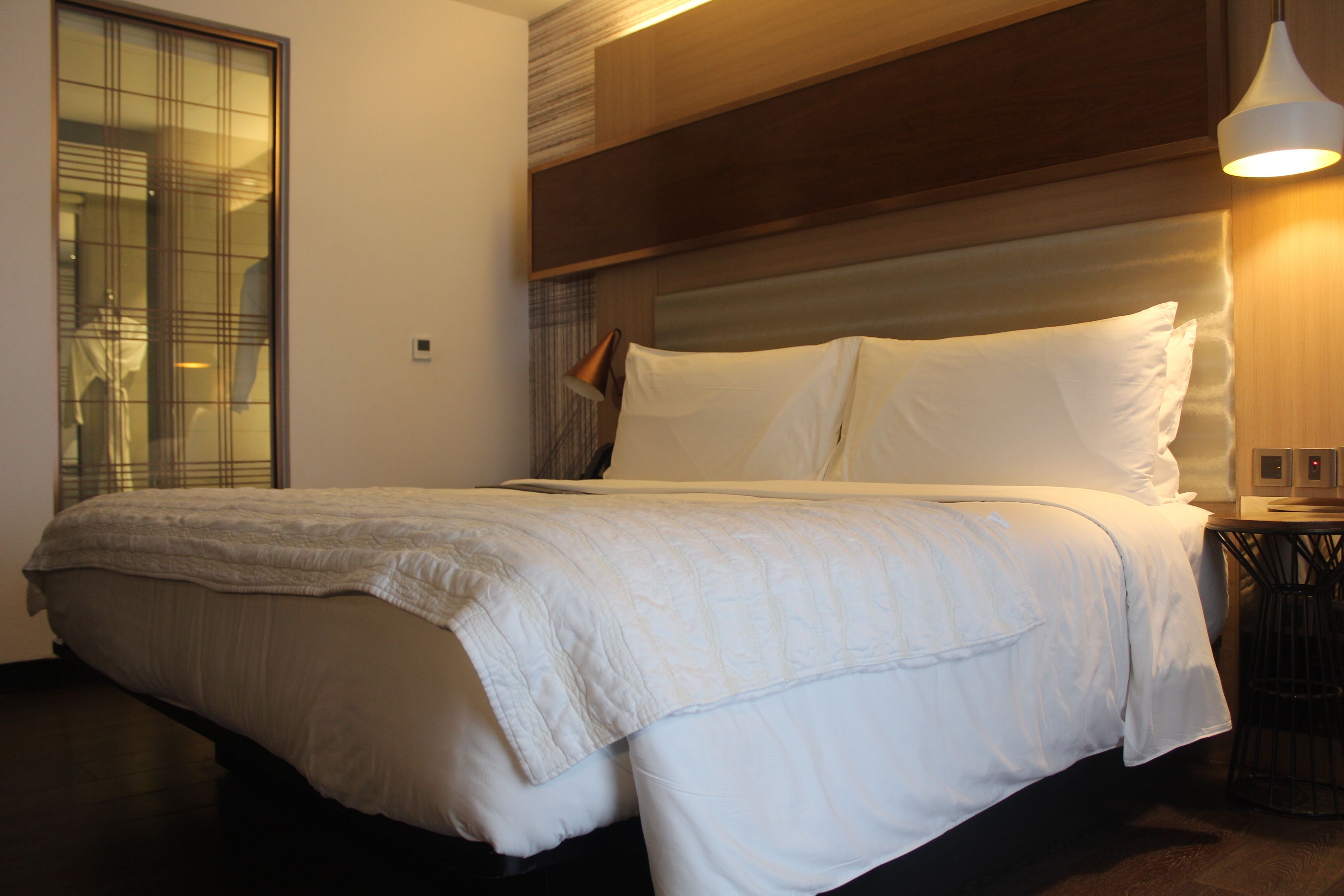 A neatly made bed with a white comforter and pillows, a wooden headboard, a bedside table with a lamp, and a view of a window with blinds.
