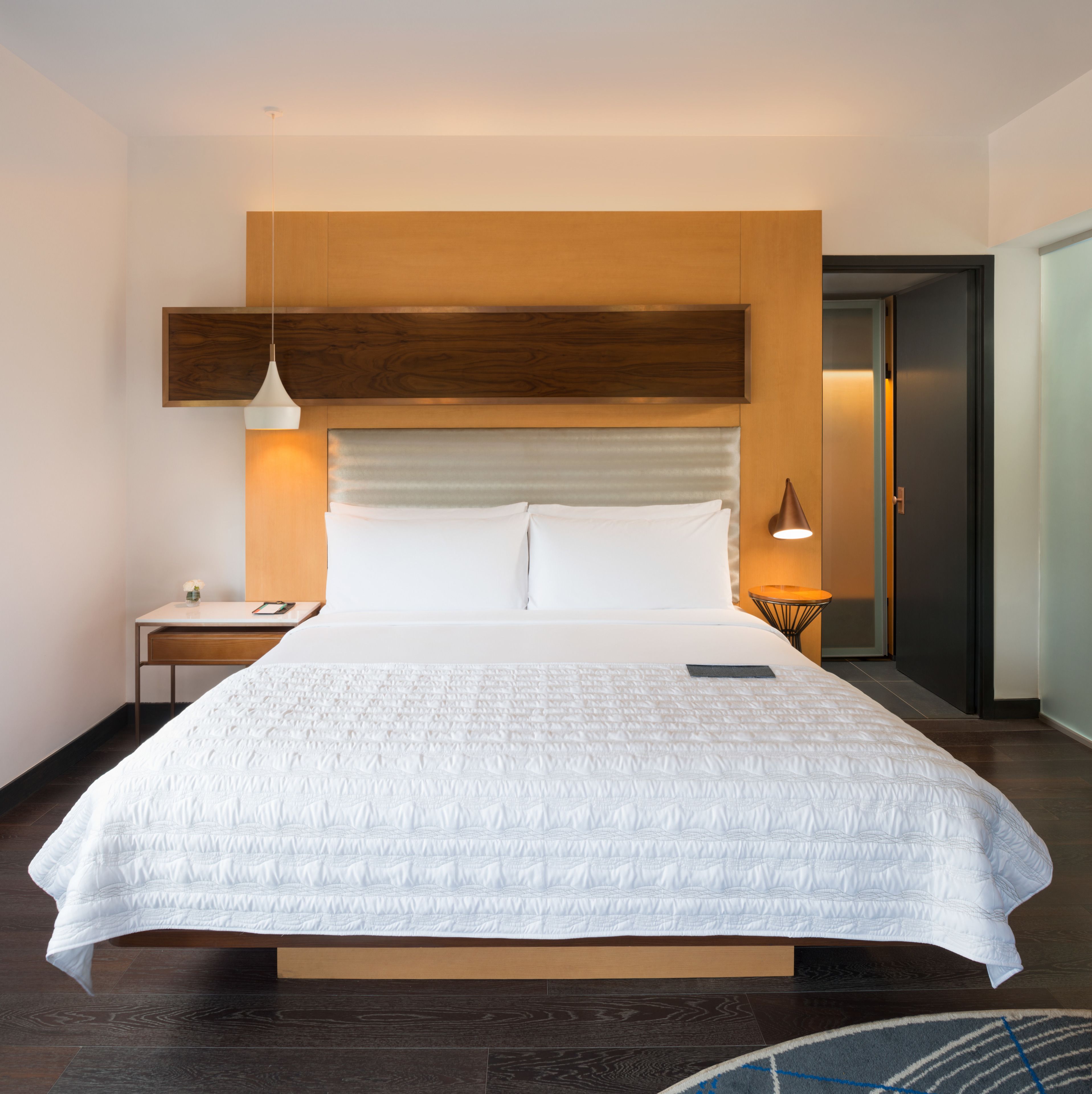 A modern hotel room with a large bed, wooden headboard, bedside tables, and a nightstand.